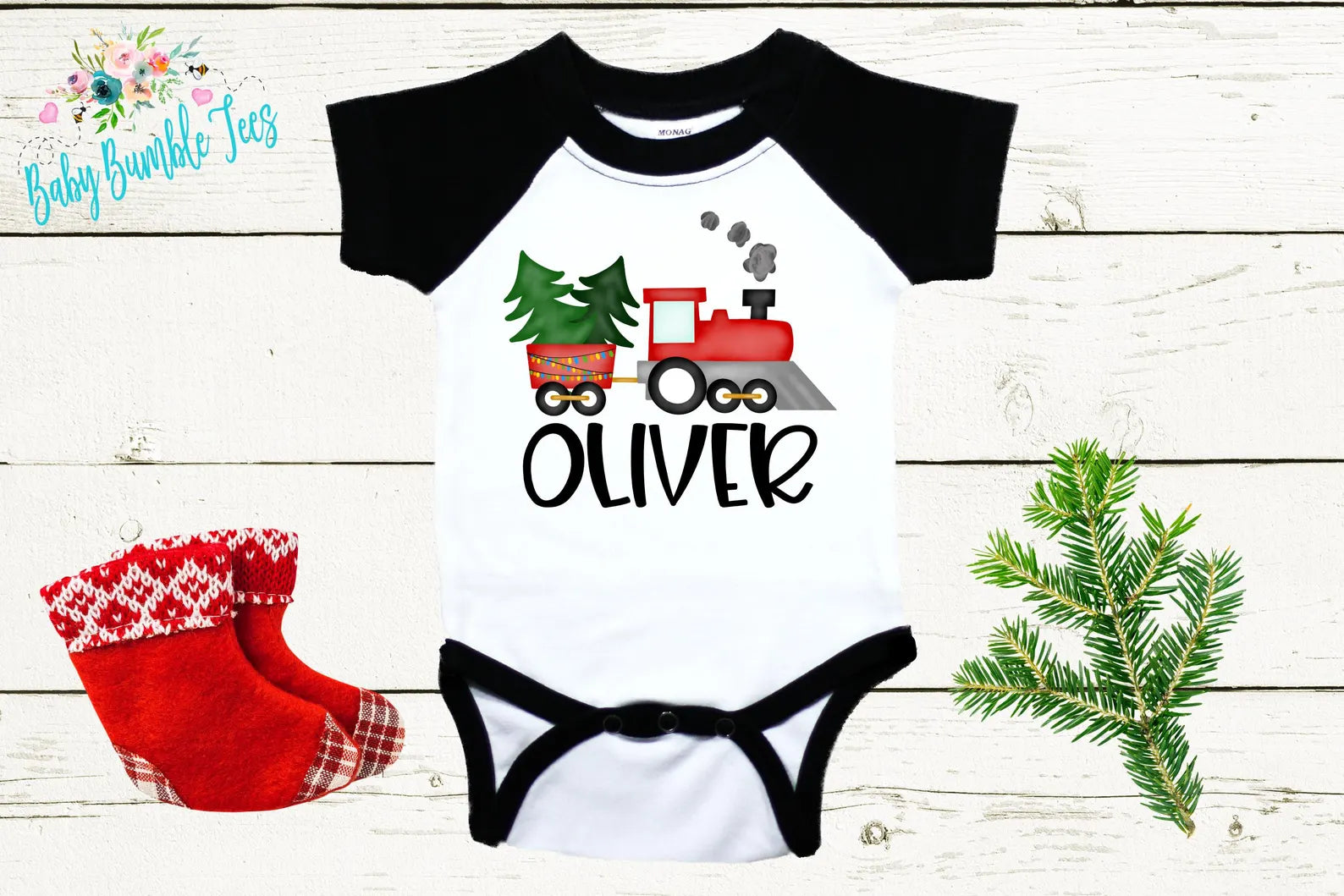 A baby onesie with a black and white design featuring a red train and a Christmas tree, along with a pair of red and white plaid socks and a pine branch.