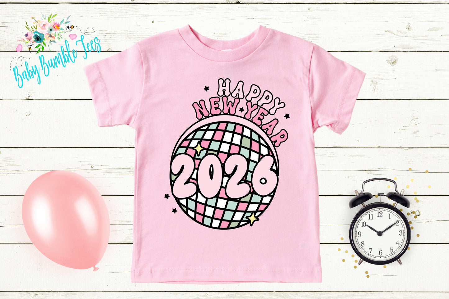 A pink t-shirt with a graphic design of a disco ball and the text "Happy New Year 2026" printed on it, along with a pink balloon and a black alarm clock on a white wooden background.