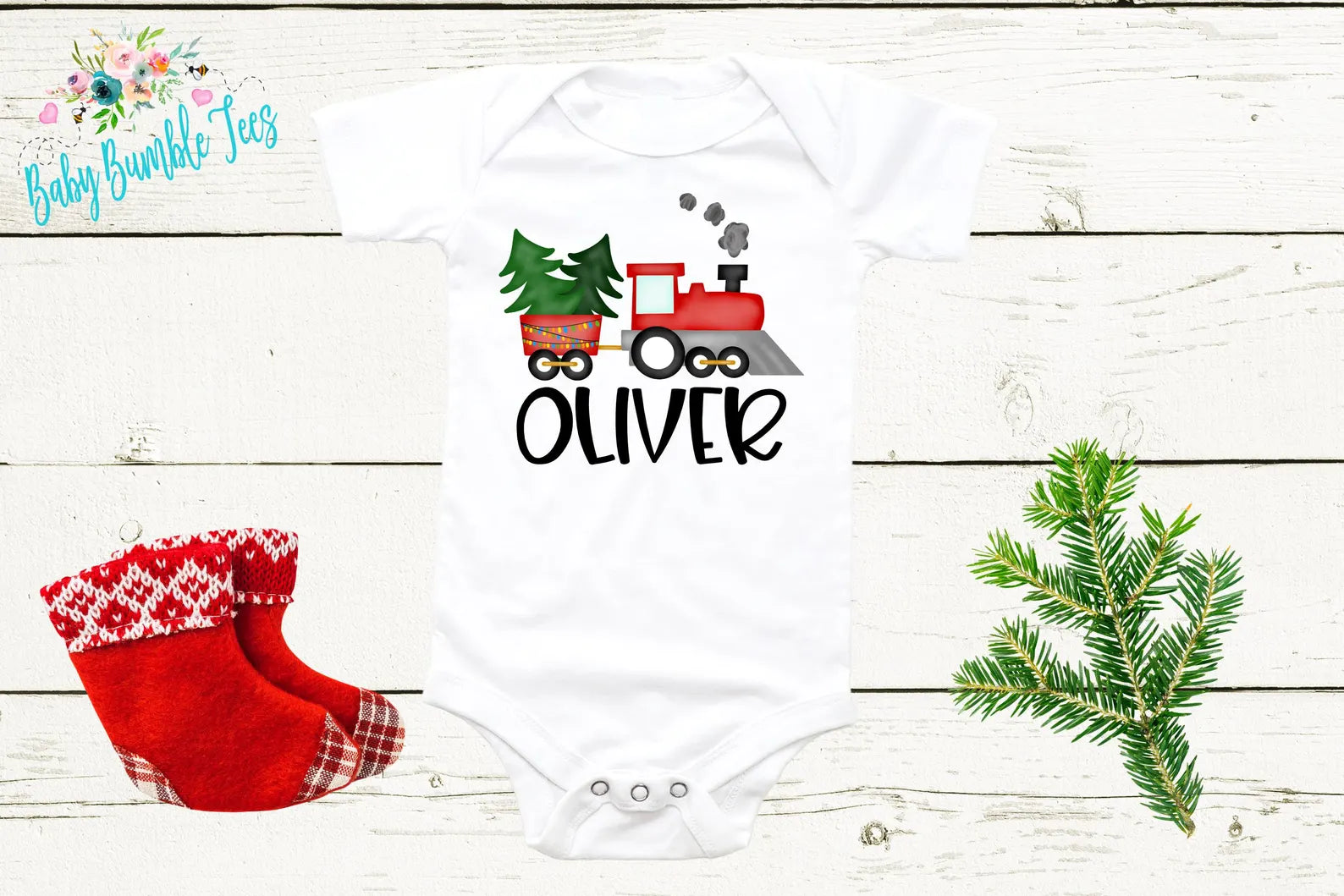A white baby onesie with a red train and the name "OLIVER" printed on it, along with a pair of red and white plaid socks and a pine branch.