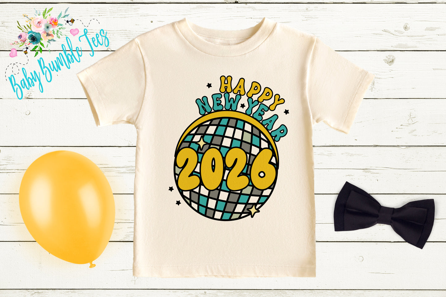 A white t-shirt with a graphic design featuring a disco ball and the text "Happy New Year 2026" is displayed on a wooden surface alongside a yellow balloon, a black bow tie, and a floral arrangement.
