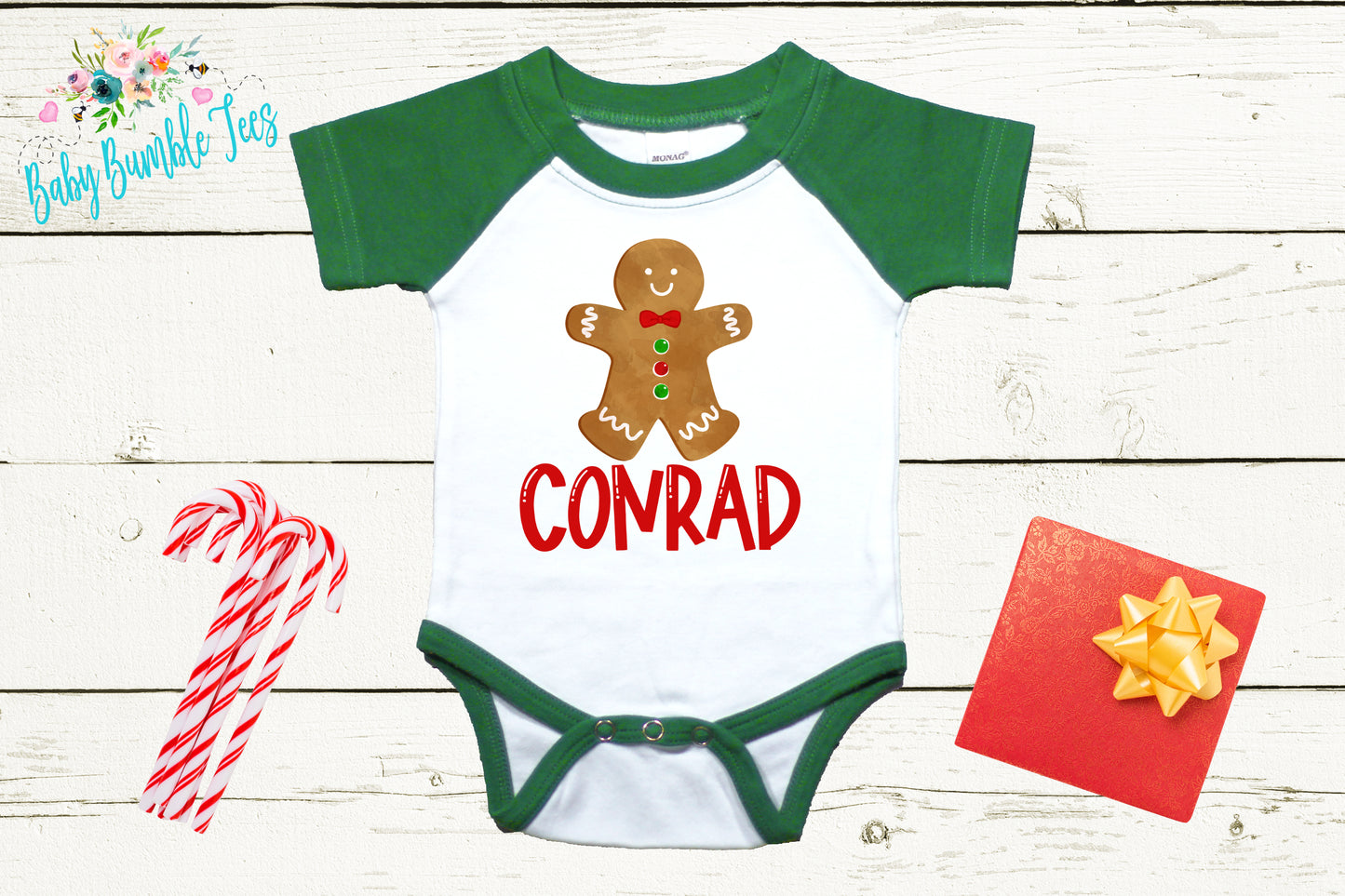 A white onesie with a green sleeve and a gingerbread man graphic, along with a red bow, candy canes, and a red gift box, all arranged on a white wooden background.