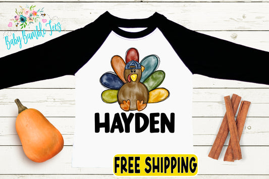 A white and black long-sleeved shirt with a colorful turkey graphic and the name "Hayden" printed on it, along with a pumpkin, cinnamon sticks, and a "FREE SHIPPING" label.