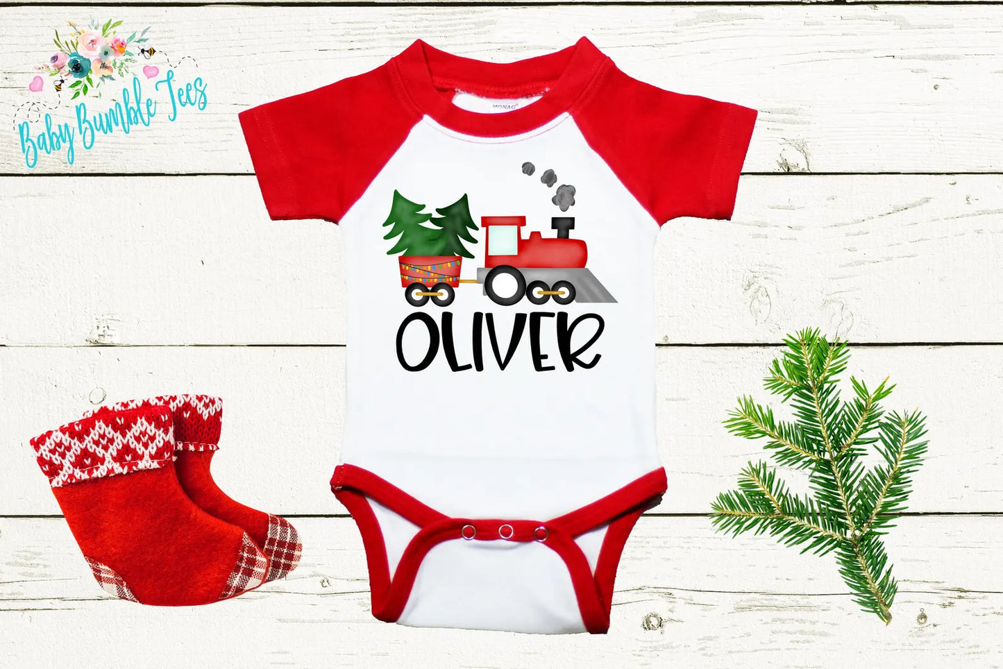 A red and white baby onesie with a train design, a pair of red and white plaid socks, and a small evergreen branch on a white wooden background.