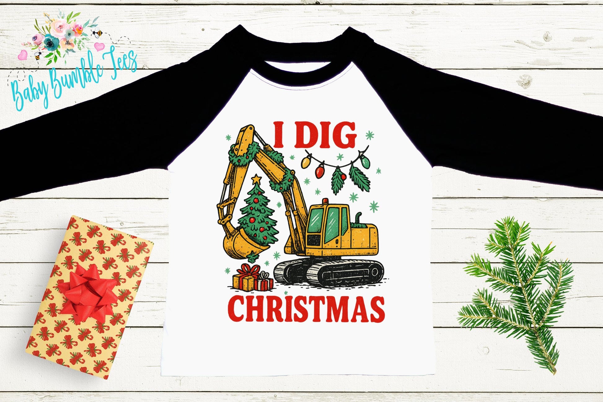 a white t-shirt with a graphic design depicting a yellow excavator digging up a Christmas tree, surrounded by holiday decorations such as lights and a wreath. The text on the t-shirt reads "I dig Christmas".