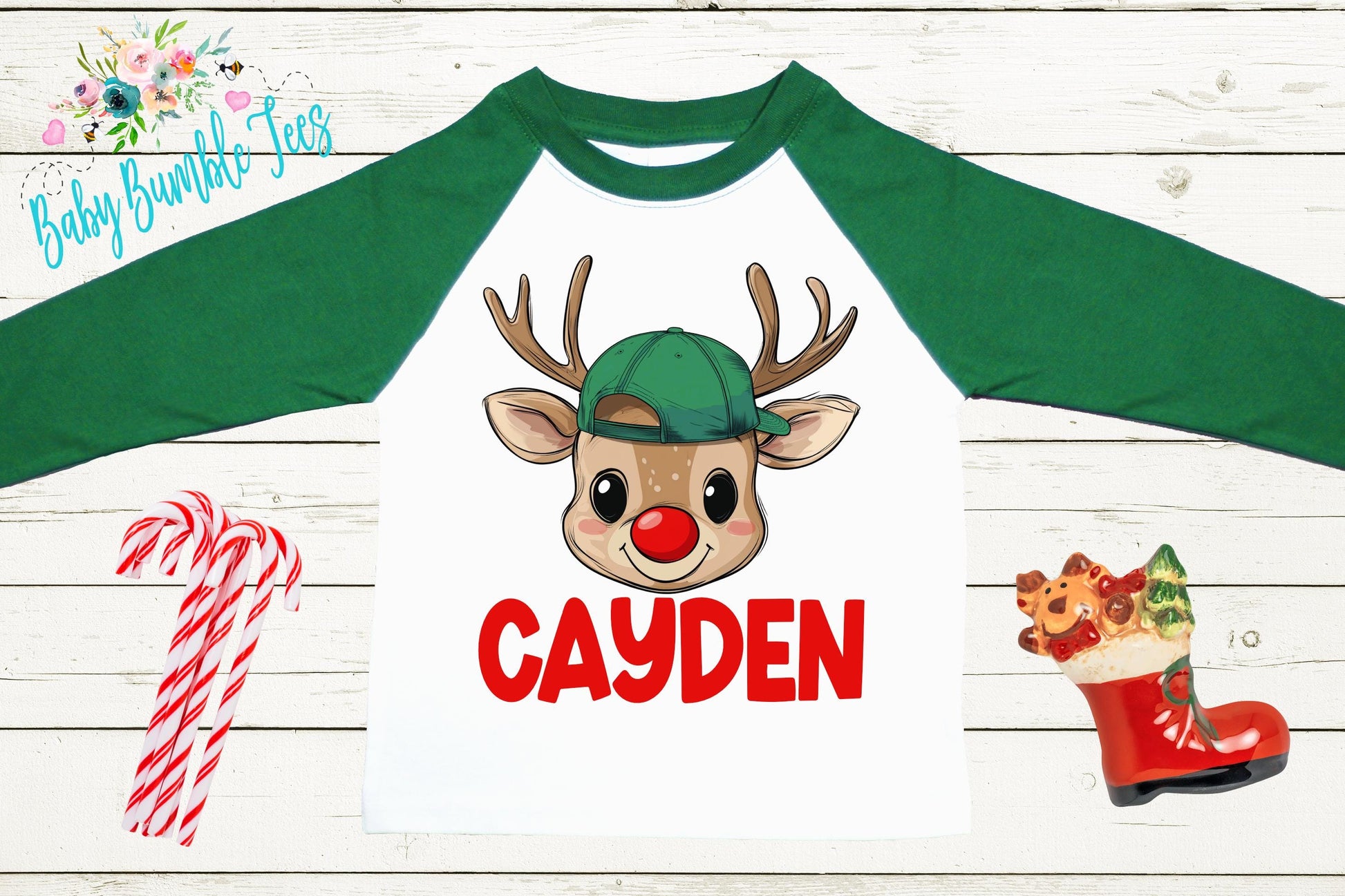 A white long-sleeve t-shirt with a cartoon reindeer wearing a green hat and the name " Cayden" printed on it. The t-shirt is displayed on a wooden surface, along with some candy canes and a small Christmas stocking.