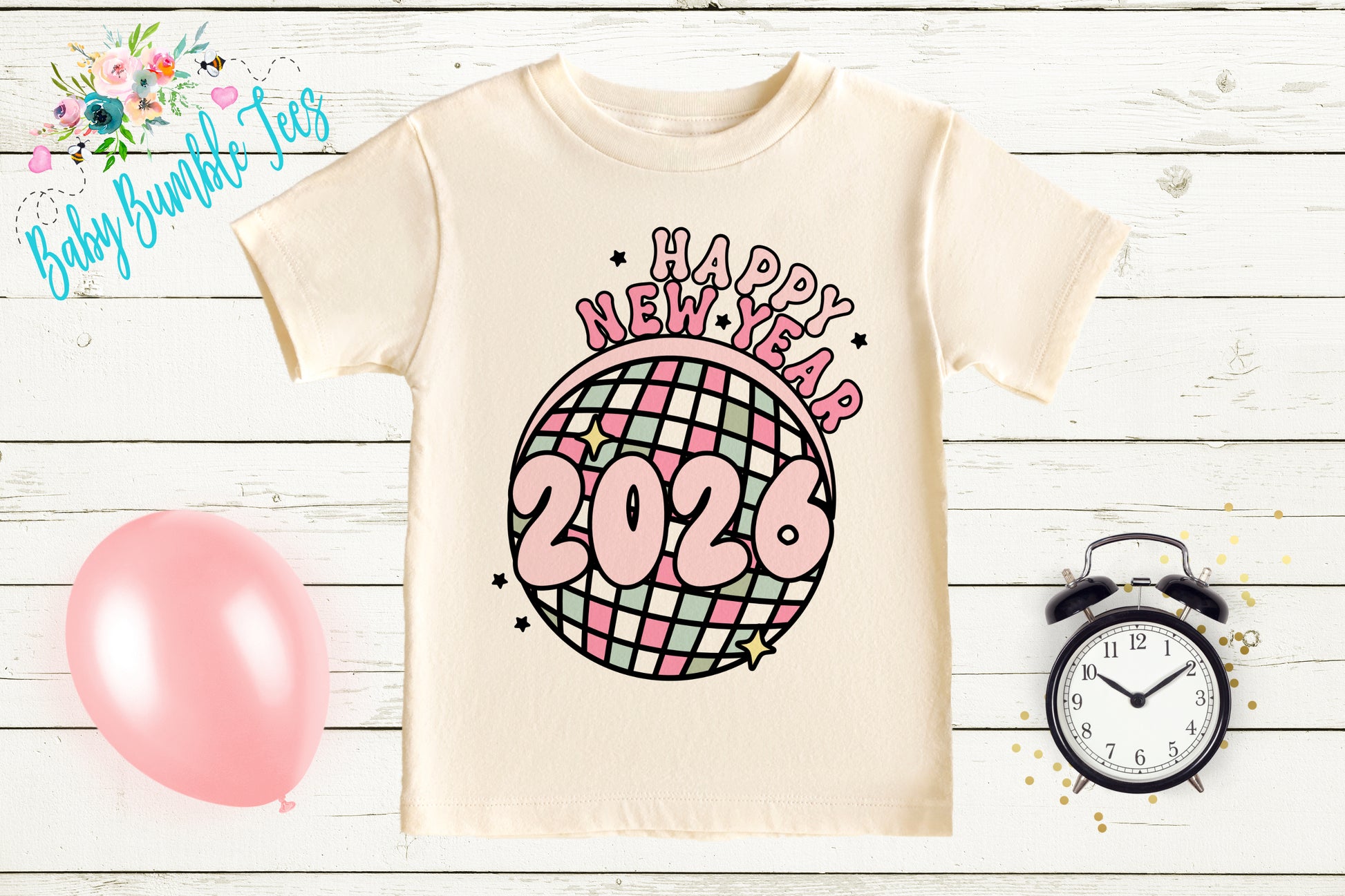 A white t-shirt with a graphic design featuring a disco ball and the text "Happy New Year 2026" is displayed on a wooden surface. Next to the t-shirt is a pink balloon, an alarm clock, and some confetti.