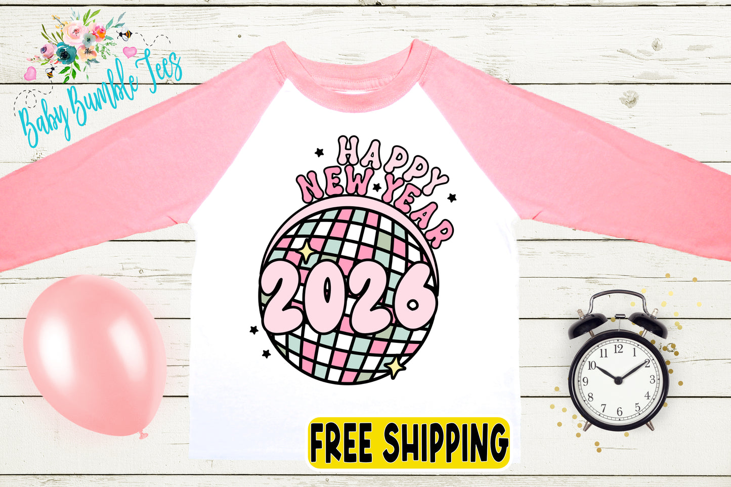 A pink and white long-sleeved shirt with a graphic design featuring a disco ball and the text "Happy New Year 2026" printed on it. The shirt is displayed on a wooden surface, along with a pink balloon, a black alarm clock, and a "FREE SHIPPING" sign.