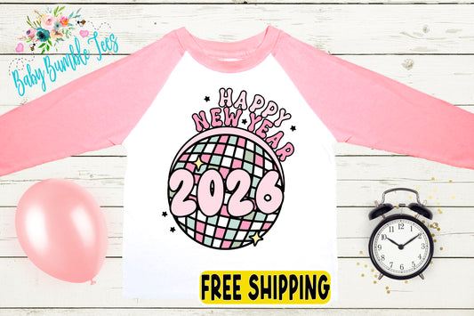 A pink and white long-sleeved shirt with a graphic design featuring a disco ball and the text "Happy New Year 2026" printed on it. The shirt is displayed on a wooden surface, along with a pink balloon, a black alarm clock, and a "FREE SHIPPING" sign.