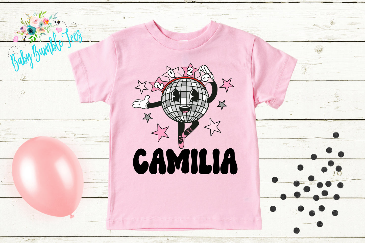 A pink t-shirt with a cartoon image of a dancing disco ball and the name "Camila" printed on it, along with a pink balloon and scattered black dots on a wooden background.