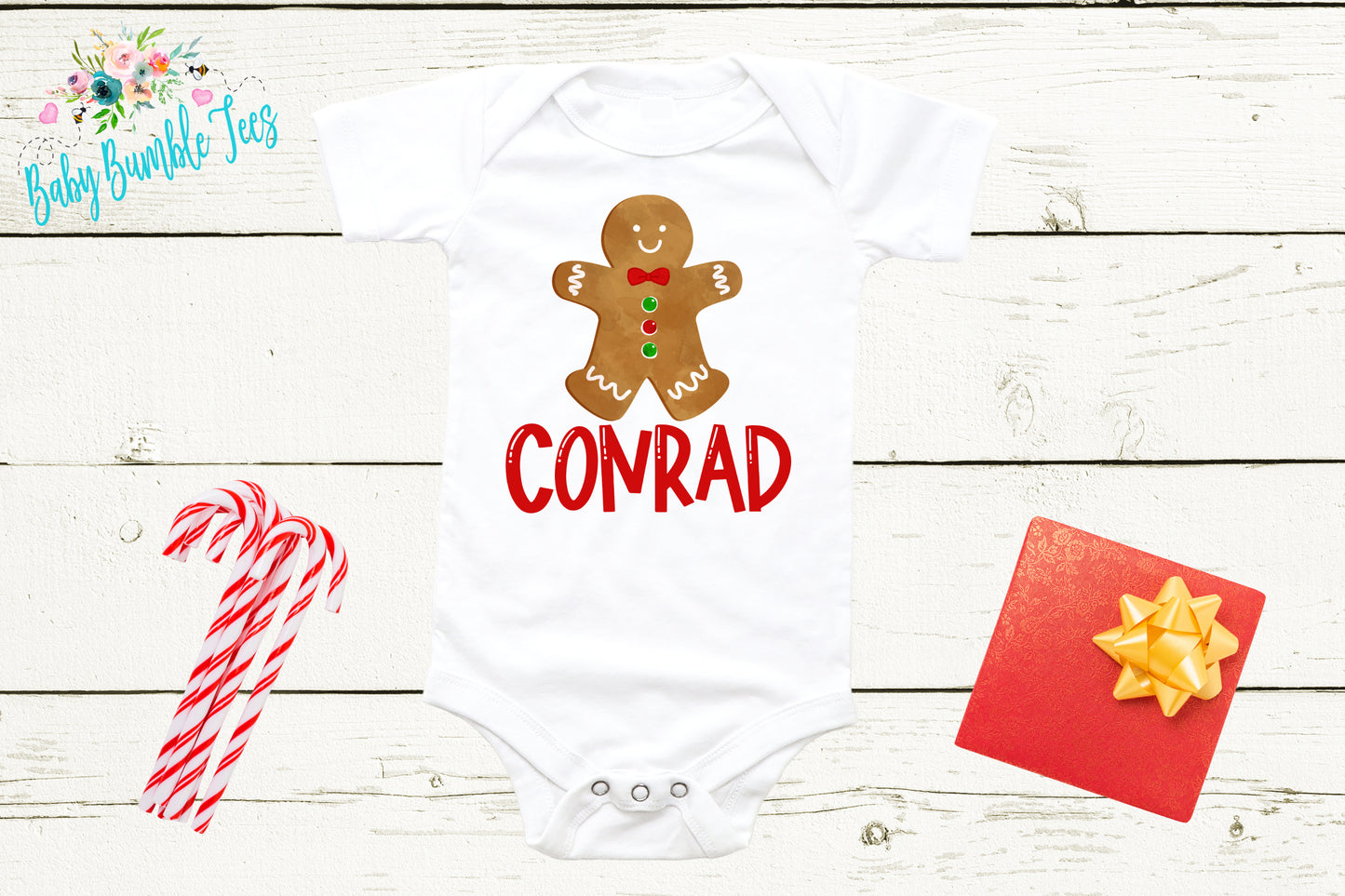 A white baby onesie with a gingerbread man design and the word "CONRAD" printed on it, along with a red bow tie, a pair of candy canes, and a red gift box with a yellow bow.