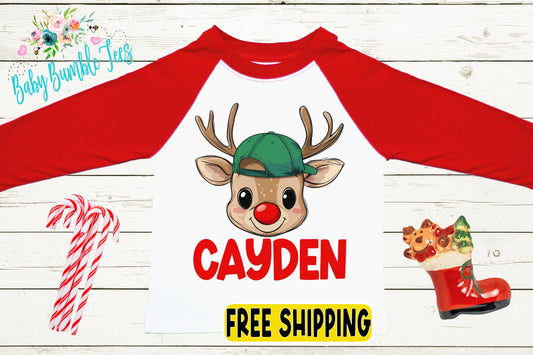 A red and white long-sleeved shirt with a cartoon reindeer wearing a green hat and the name " Cayden" printed on it. The shirt is displayed on a white wooden background, along with a pair of candy canes and a red Santa hat.