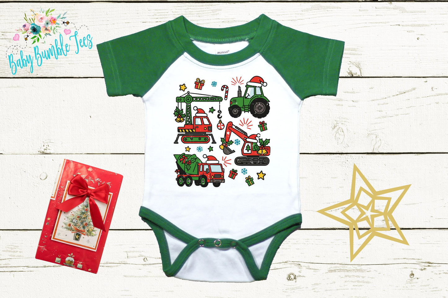 A green and white baby onesie with a graphic of construction vehicles and a Santa hat, along with a red gift box and a gold star decoration, all placed on a white wooden background.