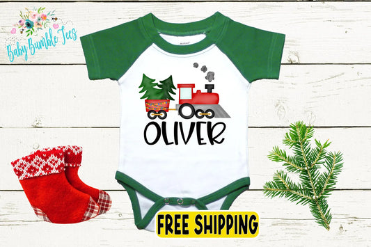 a baby onesie with a Christmas-themed design, including a train and trees, along with a pair of red and white plaid socks and a pine branch.