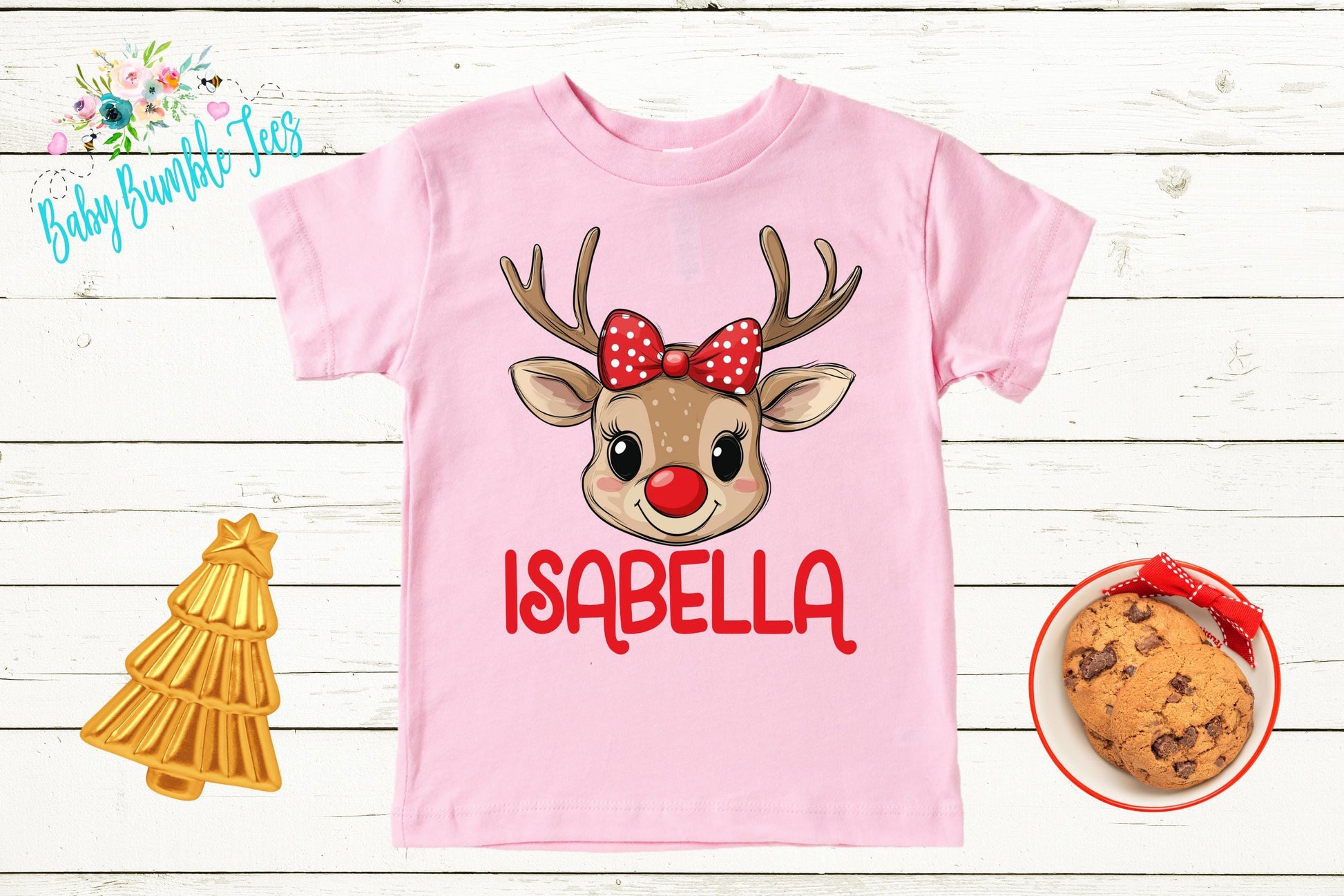 a pink t-shirt with a cartoon reindeer wearing a red bow and the name "Isabella" written on it. The t-shirt is displayed on a white wooden background, along with a small Christmas tree, a cookie, and a cookie-shaped ornament.