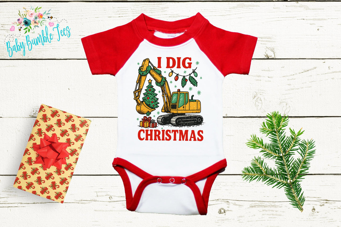 A red and white baby onesie with a graphic of a construction vehicle and the text "I dig christmas" printed on it, along with a Christmas tree and a gift box.