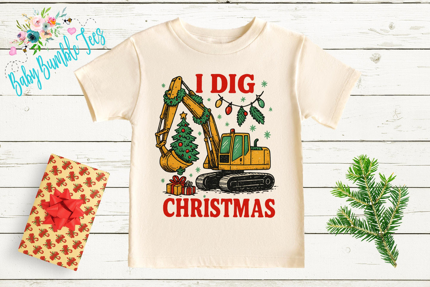 a white t-shirt with a graphic design depicting a yellow excavator digging a Christmas tree, surrounded by Christmas decorations and gifts.