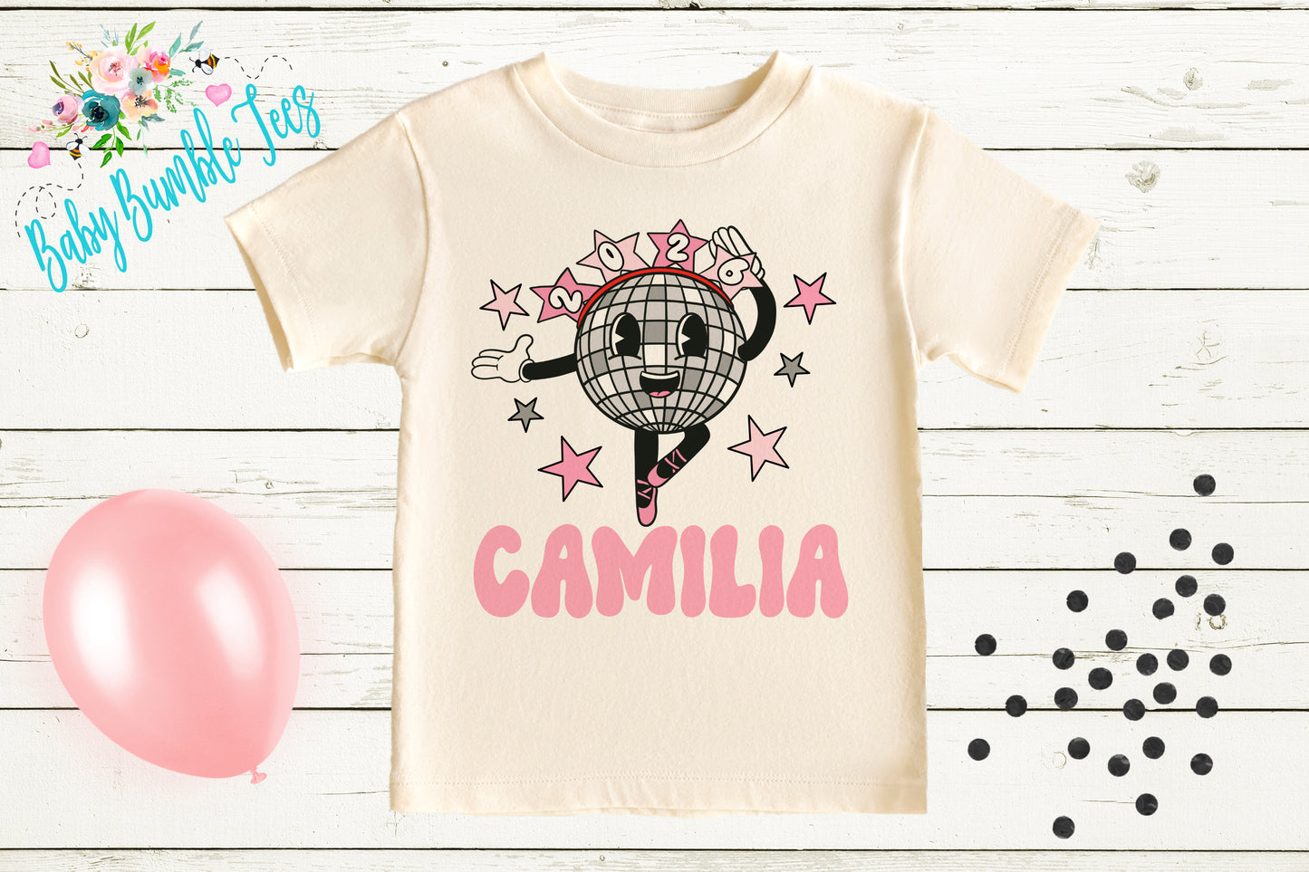 a white t-shirt with a cartoon character and the name "CAMILIA" printed on it, along with a pink balloon and scattered black dots on a wooden background.