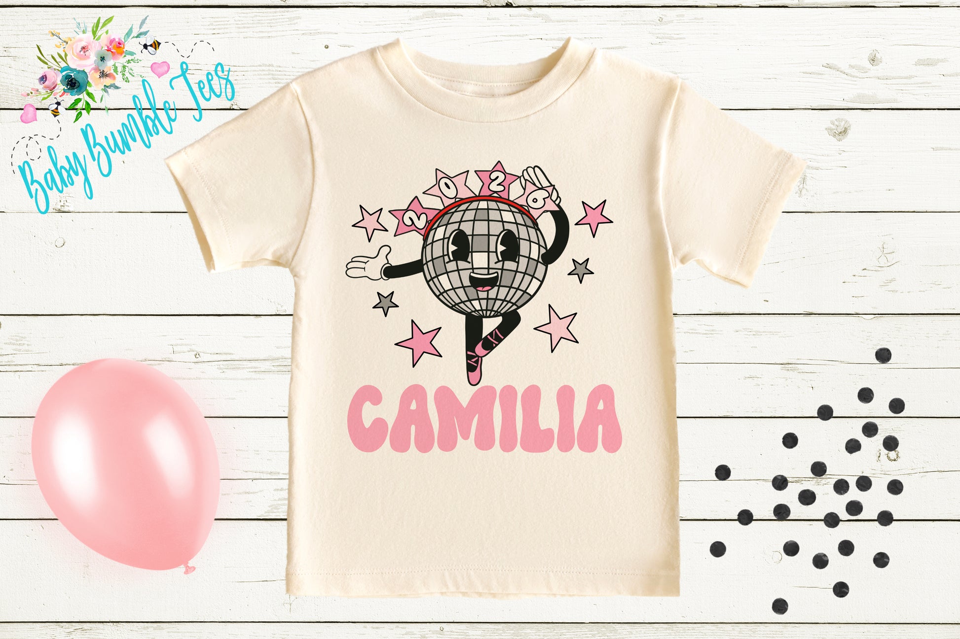 a white t-shirt with a cartoon character and the name "CAMILIA" printed on it, along with a pink balloon and scattered black dots on a wooden background.