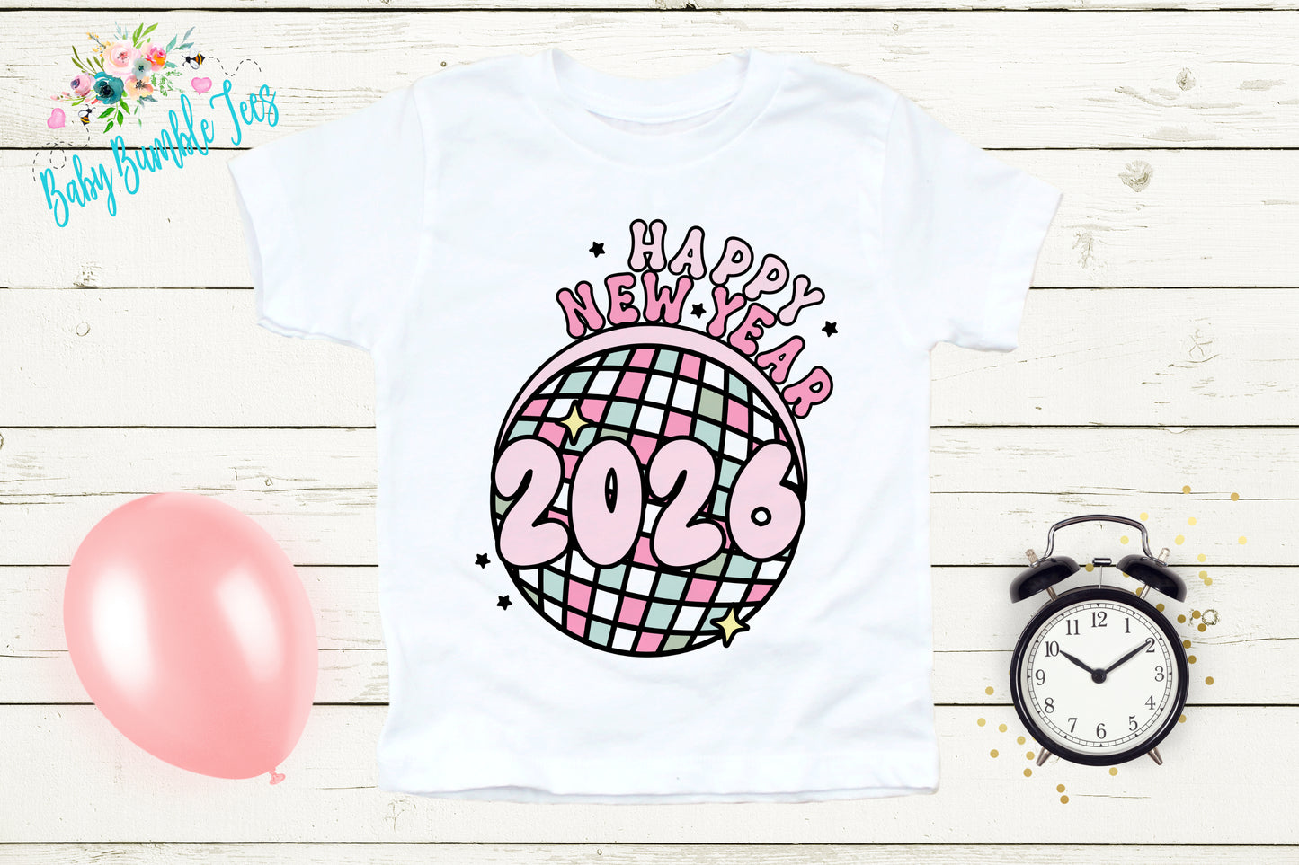 A white t-shirt with a graphic design featuring a disco ball and the text "Happy New Year 2026" is displayed on a wooden surface. Next to the t-shirt are a pink balloon, a black alarm clock, and some confetti.