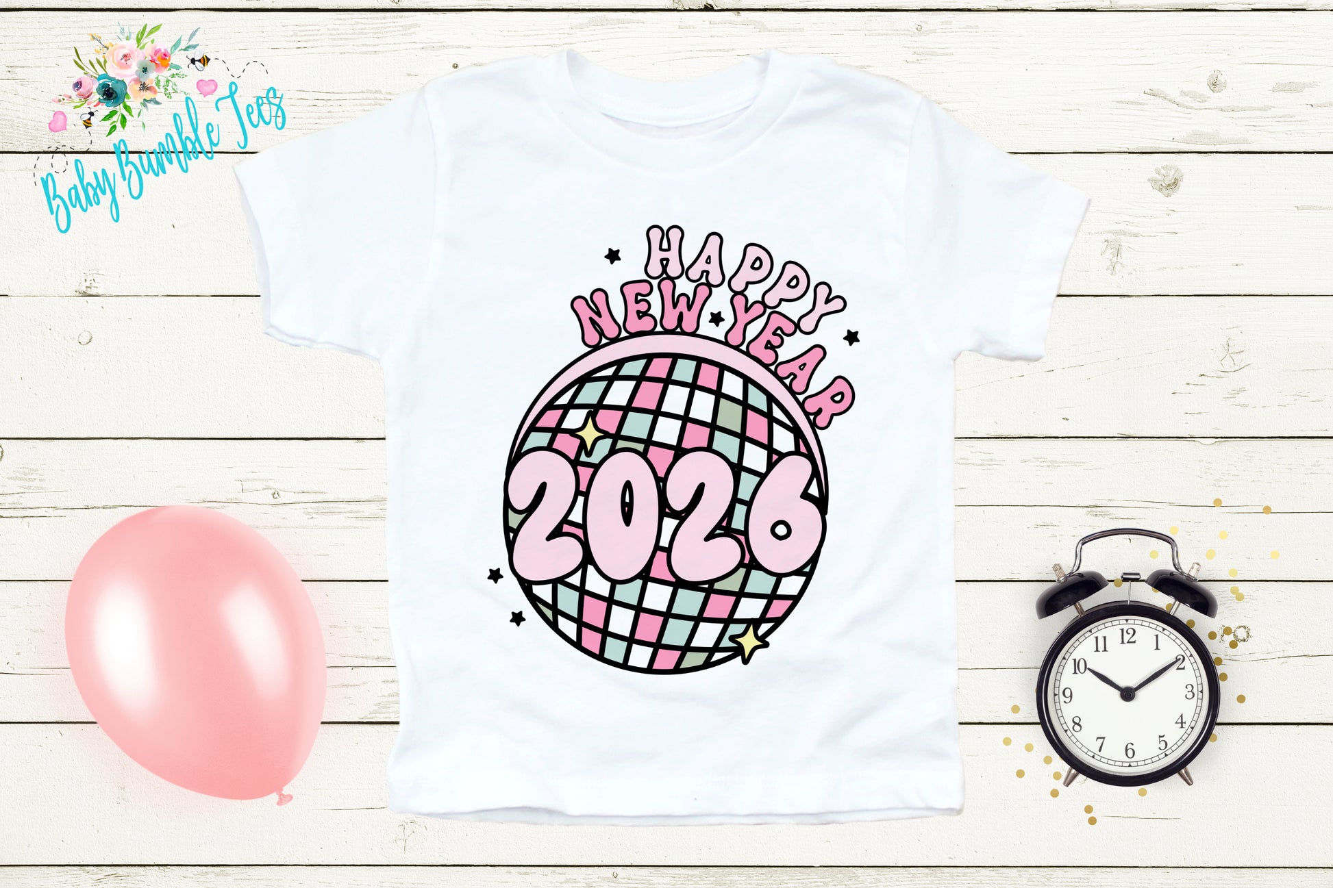 A white t-shirt with a graphic design featuring a disco ball and the text "Happy New Year 2026" is displayed on a wooden surface. Next to the t-shirt are a pink balloon, a black alarm clock, and some confetti.