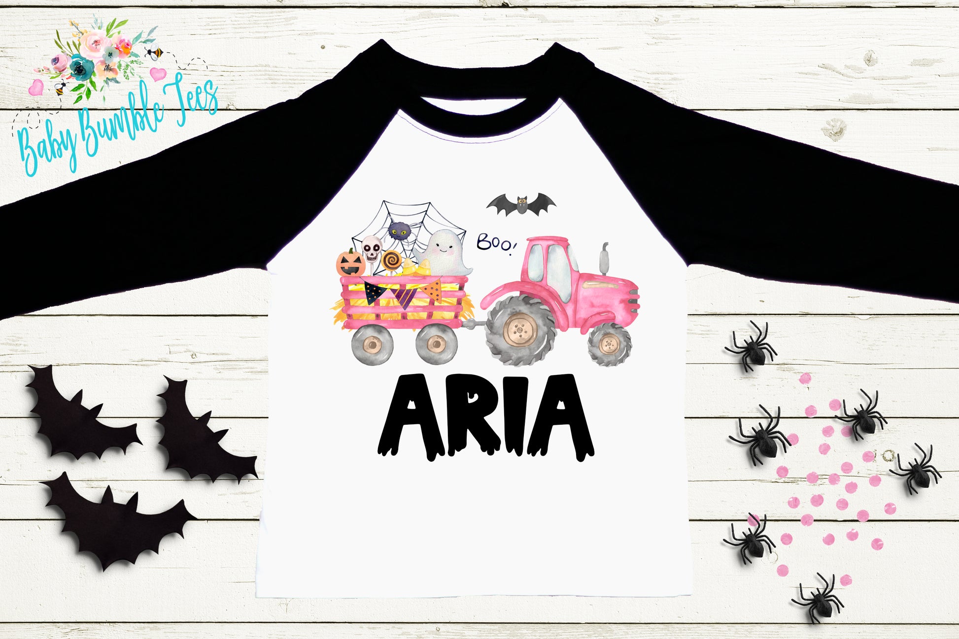 a white long-sleeved shirt with a graphic design of a pink tractor and various animals, including bats and a cat, on the front. The shirt has the name "ARIA" printed on it. The shirt is displayed on a wooden surface, surrounded by black bats and pink and black spider decorations.