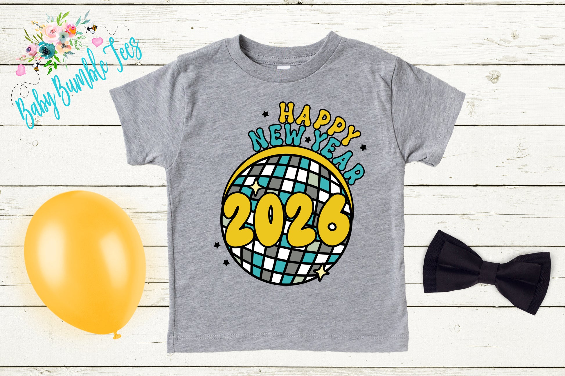 A gray t-shirt with a graphic design featuring a disco ball and the text "Happy New Year 2026" is displayed on a wooden surface alongside a yellow balloon, a black bow tie, and a floral arrangement.