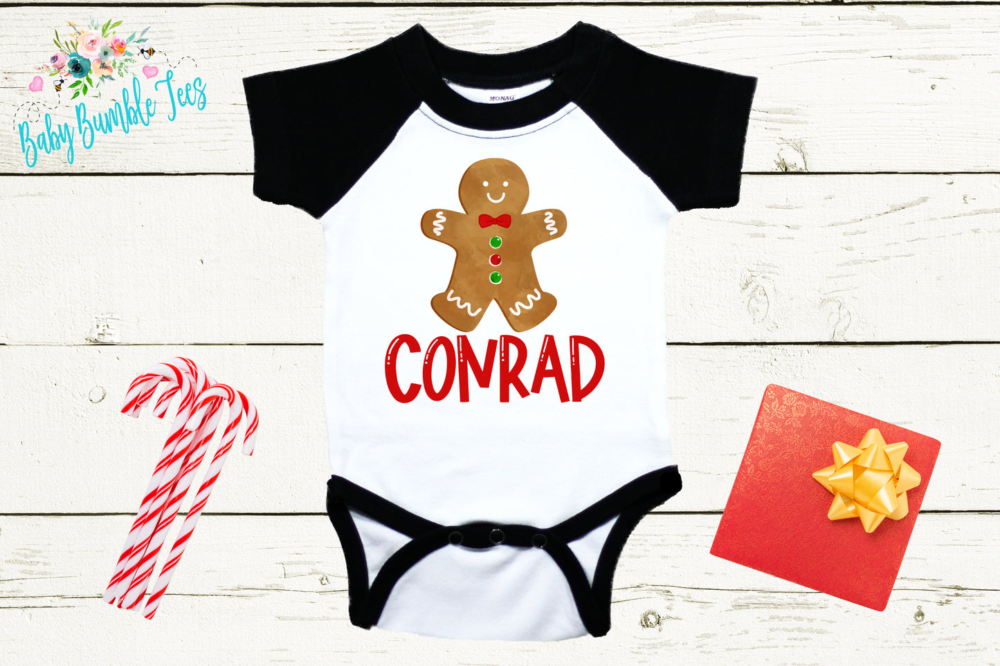 A white baby onesie with a black and red design, featuring a gingerbread man and the word "CONRAD" printed on it. The onesie is displayed on a white wooden background, accompanied by a pair of candy canes, a red gift box, and a yellow bow.