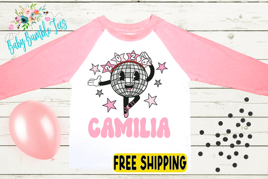 a pink and white long-sleeved shirt with a cartoon image of a disco ball and stars, along with the name "Camila" printed on it. The shirt is displayed on a wooden surface, and there are pink balloons and black dots around it.