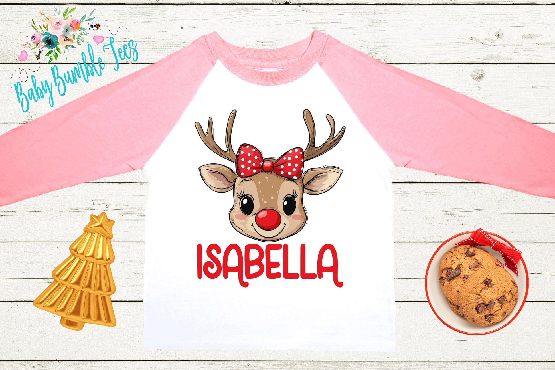 A pink and white long-sleeved shirt with a cartoon reindeer wearing a red bow and the name "Isabella" printed on it. The shirt is displayed on a wooden surface, along with a small Christmas tree, a cookie, and a plate of cookies.