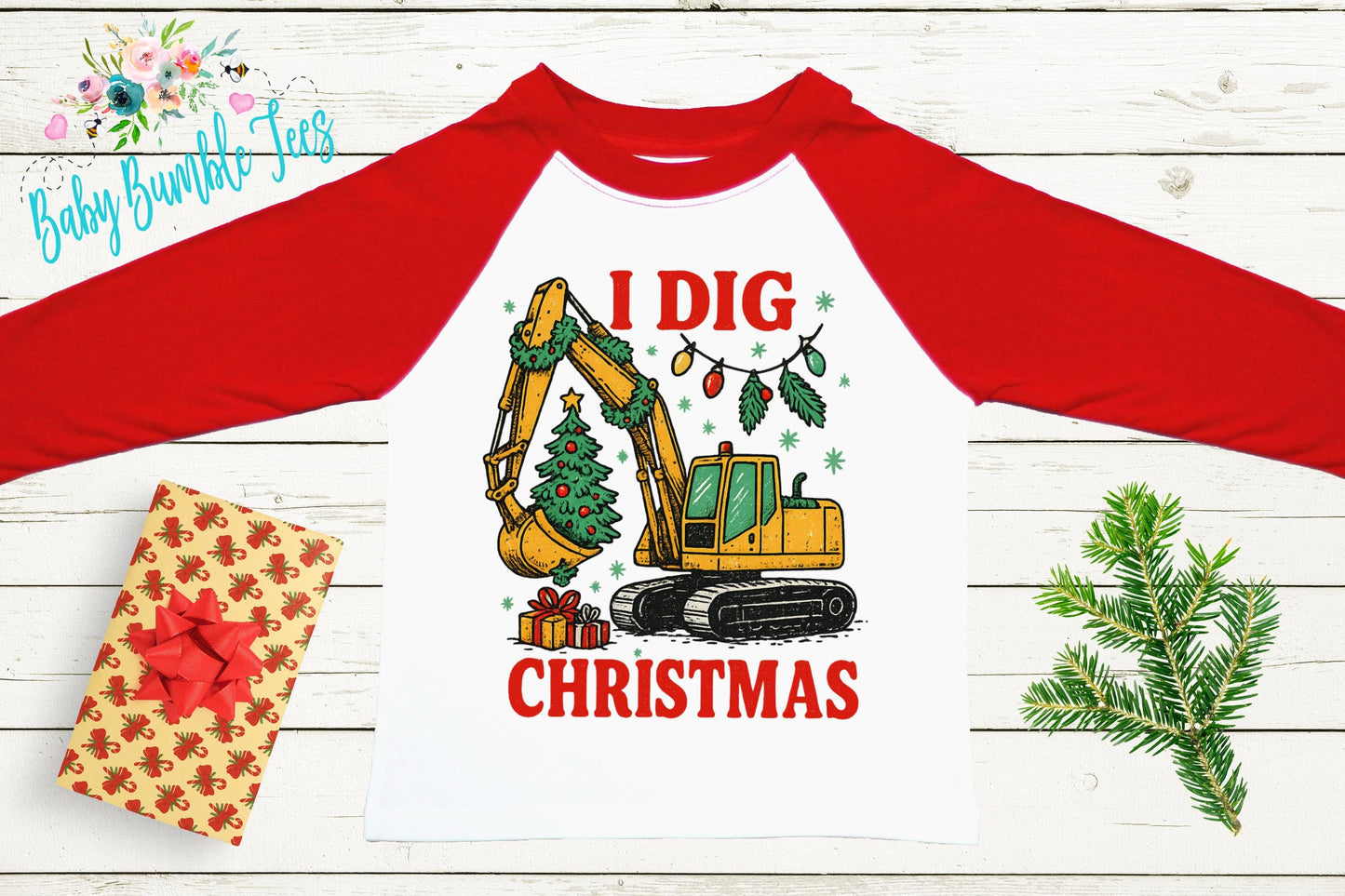 a red and white long-sleeved shirt with a graphic design depicting a yellow excavator digging a Christmas tree. The shirt has the text "I dig Christmas" printed on it. The shirt is placed on a white wooden surface, with a gift box, pine branches, and a Christmas tree decoration nearby.